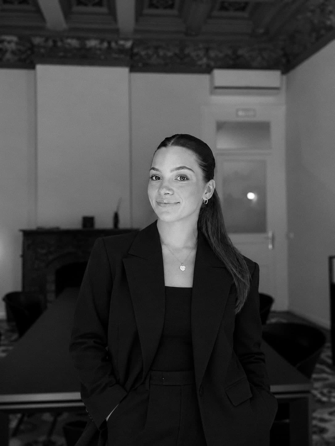 Black-and-white portrait of Rebekah Guasto in a black blazer, smiling, in a room with an ornate ceiling